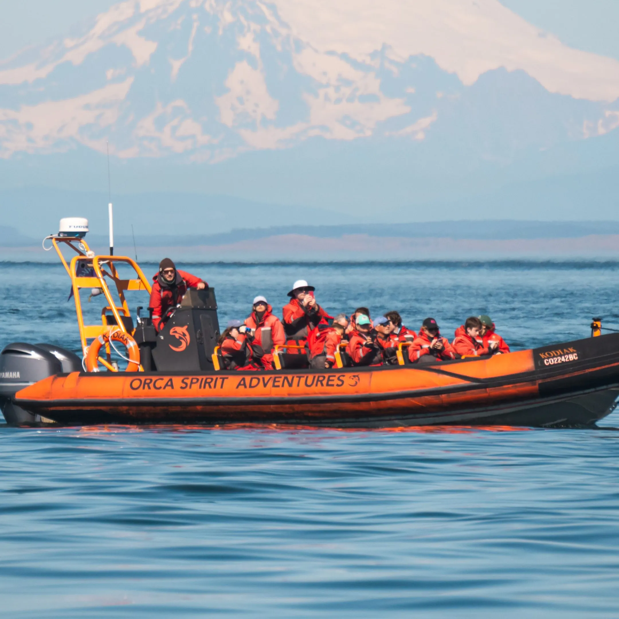 Orange inflatable boat with people in life jackets on calm water, snow-capped mountain in the background.