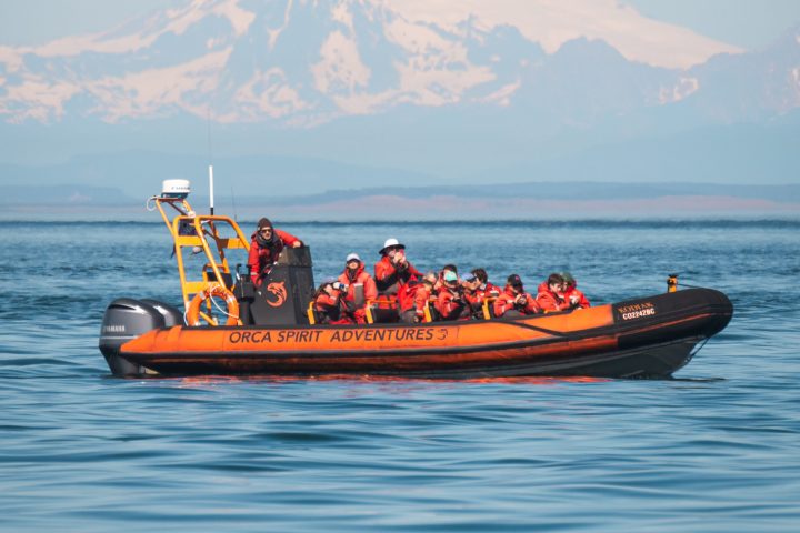 Orange inflatable boat with people in life jackets on calm water, snow-capped mountain in the background.