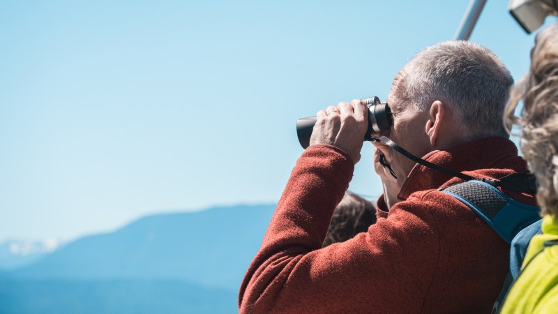 Person in red jacket using binoculars to view distant mountain landscape.