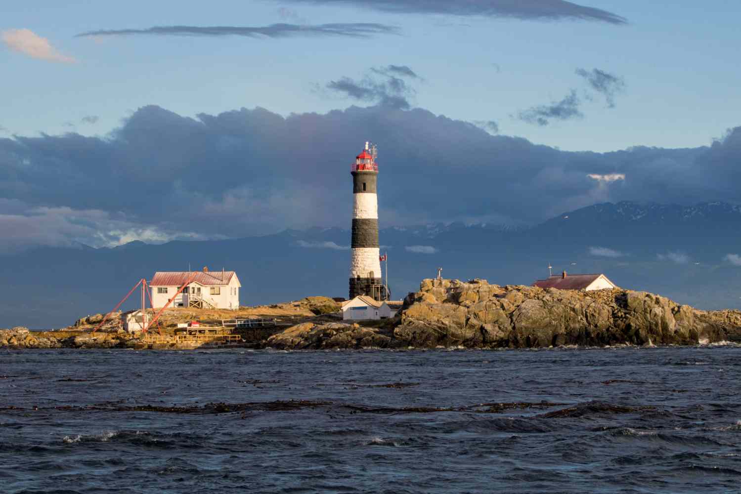 Lighthouse and buildings on rocky island with cloudy sky and ocean in foreground.