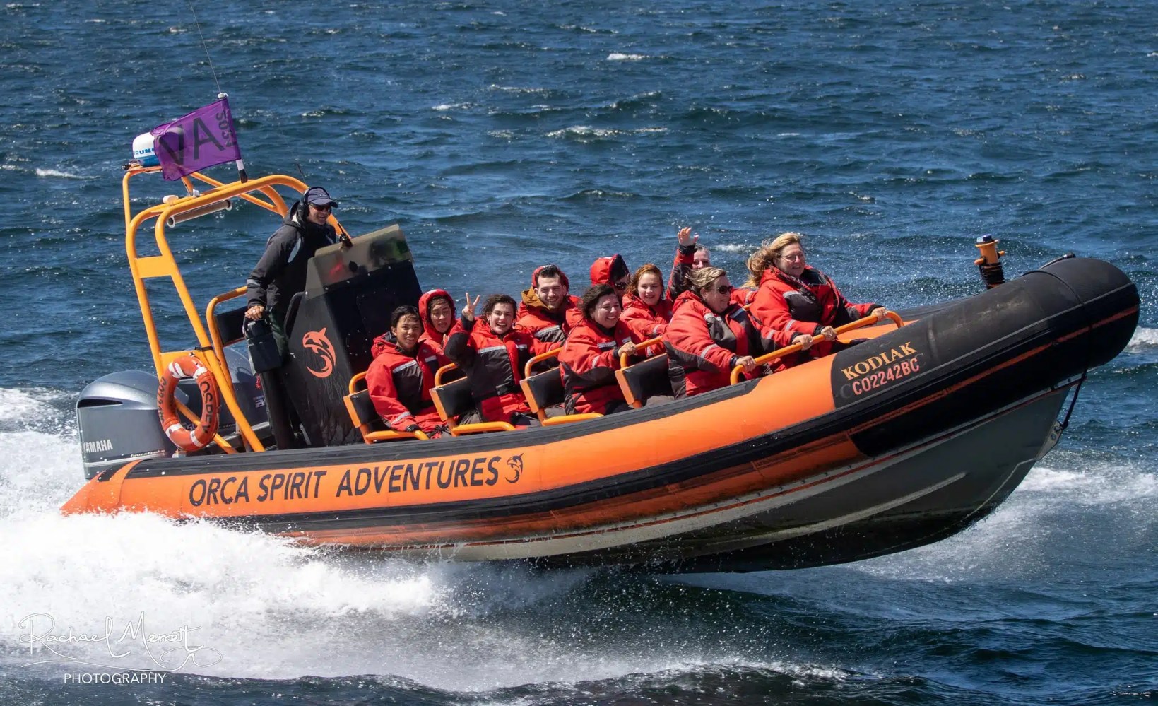 People in orange jackets on a zodiac boat named 'Orca Spirit Adventures' speeding in the ocean.