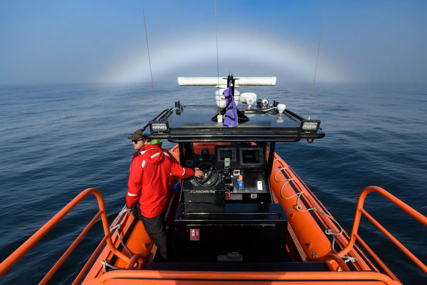 Person on orange boat navigating under a fogbow at sea.