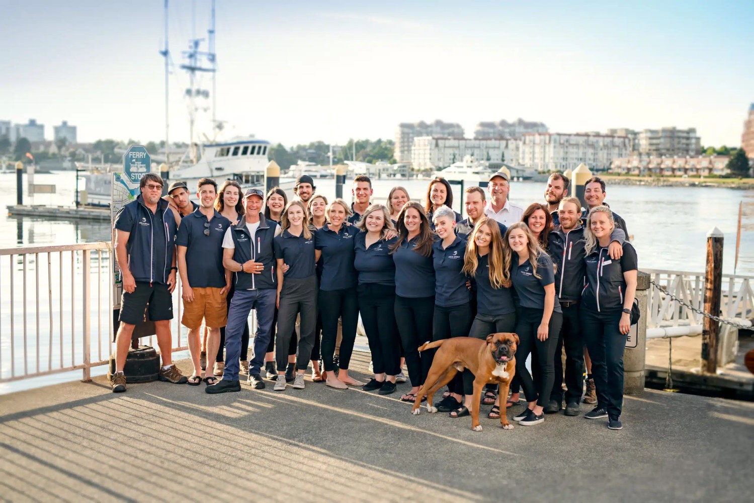 Group of people and a dog posing outdoors by a pier with boats in the background.