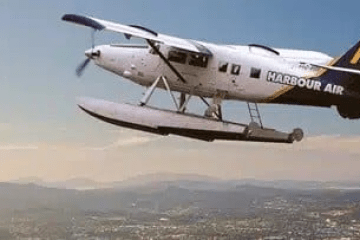 A seaplane flying over a cityscape with mountains in the background.